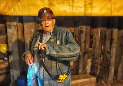 An older man in a hard hat and work coveralls appears as if he is speaking in a highly industrial location