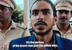 A still from a movie. A dark-skinned man, dressed in a blue shirt, is flanked on either side by men dressed in military garb. The text (which is a subtitle) reads, "It is the century of the brown man and the yellow man."