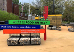 A photograph of an outdoor art installation. In the foreground, an oversized fork stands besides signs that read, "What is food justice? We all makes decisions about what we eat...but we don't make those decisions by ourselves.”