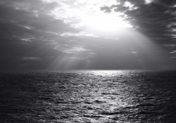 A black and white photograph of the sun peeking through the clouds above the ocean