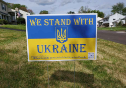 A photograph of a sign in a yard in a residential neighborhood. Text reads: We stand with Ukraine. 100% of all proceeds will go to the Ukrainian people.