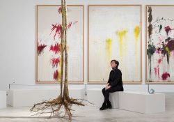 A woman is seated on a stark white bench in front of a triptych of three abstract painting while looking at a tree.