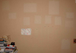 A brown wall with faded spots where pieces of paper where once affixed but have now been removed. A small pile of books and notebooks sits on a table just to the left of the frame