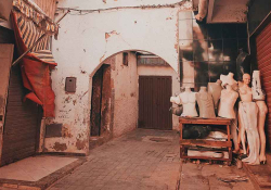 A photograph of the interior of city alley. Here, a collection of used mannequins gather outside of a stone archway