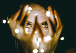 A photograph of a woman holding a string of lights in her hands, which are in front of her face. Only her eyes can be seen clearly through her splayed fingers