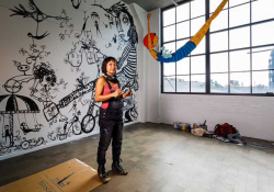 A photograph of Denise Duong standing in an art studio. There are large figures painted on the white wall behind her and the room is lit by large windows on the right.