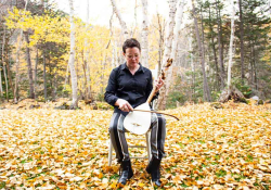 A photograph of Stephanie McKenzie, sitting in an armless chair in a natural setting, while holding a bowed instrument upright in the lap