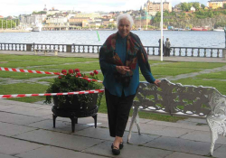 A photograph of an older woman standing by a bench, which is itself beside a river