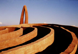 A photograph of an outdoor installation. This detail shows one edge of a series of low, concentric walls, the interiors in shadow, with a tall arc standing in the background