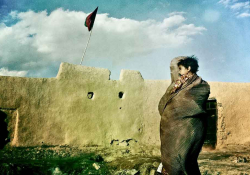 A woman veiled from head to toe, holding a child, as she looks at a stone wall with a flag flying above