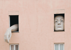 A photograph of a two windows on a two story apartment building. A face (oversized) fills one and a giant hand emerges from the other.