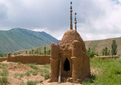A tomb, built from clay, with rounded shapes suggestive of a mosque, sits in a sparsely covered grass field outside the walls of a city