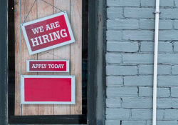 A photograph of a sign hanging in a window. Text reads; We are Hiring. Apply Today.