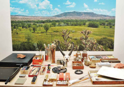 A table full of painting tools with a painting of a New Mexican landscape mounted on the wall behind it