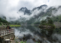 Spectators stand on a stone overlook that juts on to a wooded lake that sits at the foot of foggy mountains