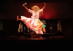 A dancer, clothed in a full length housedress, her face obscured, leaps across the stage