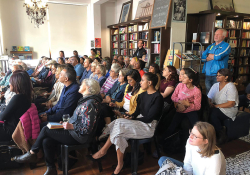 A photograph of an attentive audience, mostly seated in chairs, attending a reading in a bookstore