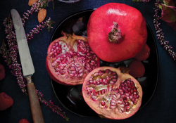 Looking down on a table that has two pomegranates on it, one cut, one uncut, with the knife that opened the one up laying beside it.