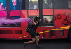A woman masked in a black bandana runs alongside a bus covered with graffiti carrying a purple flare