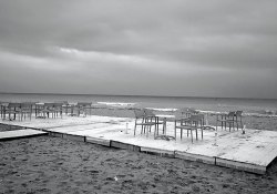 A black and white photos of empty tables and chairs on a patio overlooking a restless sea