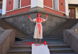 A photograph of a woman dressed in white standing at the intersection of two sets of stairs. She is splattered with red paint.