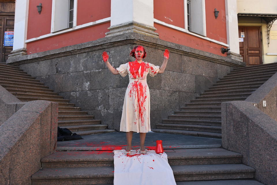 A photograph of a woman dressed in white standing at the intersection of two sets of stairs. She is splattered with red paint.