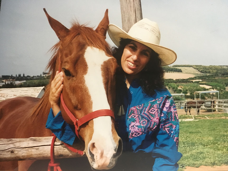 A color photograph of Margarita, wearing a cowboy hat, nuzzling a horse