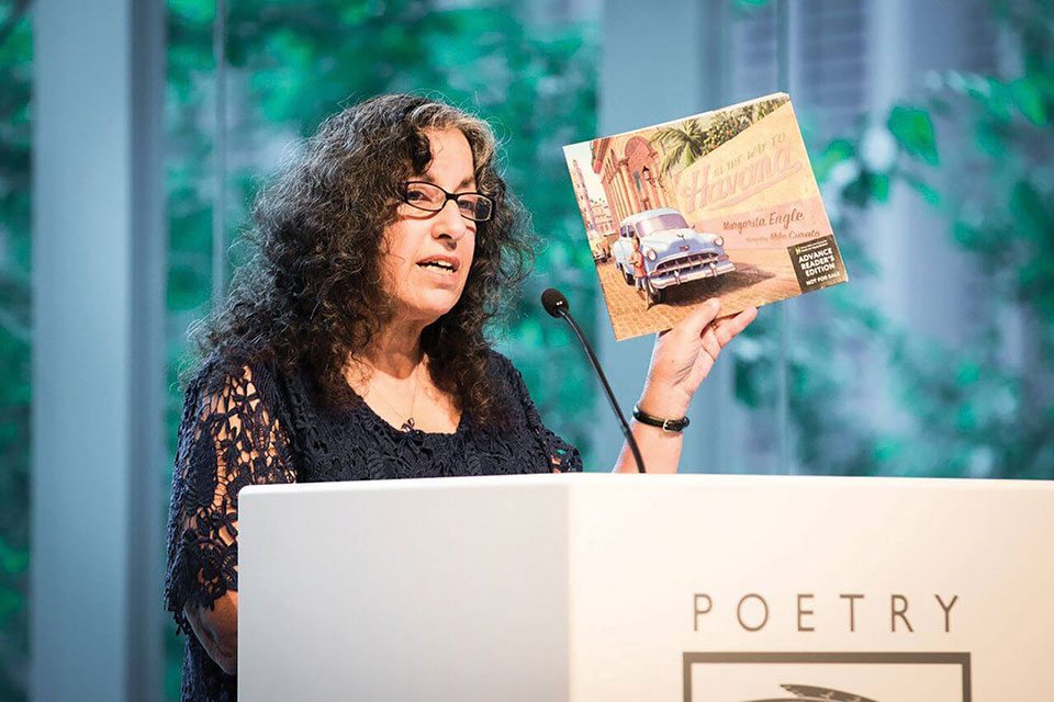 Margarita stands behind a lectern branded by the Poetry Foundation holding a copy of her book, Havana