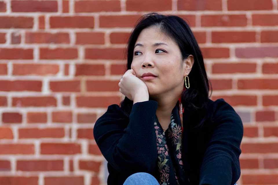 A photograph of Nicole Chung sitting in front of a red brick wall. She is looking off into the distance. 