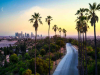 A photograph of LA at dusk. A paved road winds through tree dotted hilly landscape