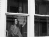 A black and white photograph of a young couple, seen from outside somberly looking at the photographer out a bay window