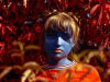A photograph of a young woman, painted blue, centered in the frame against a oversaturated field of red leaves