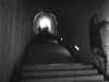 A grainy black and white photo of a stairway inside of a castle. Figures up the stairs, some distance away, are emerging into the light outside