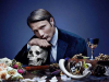 A promotional still of an actor, seated at a table, resting his chin atop his hand, which rests on a human skull