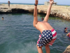 A figure clad in swimming shorts with an American flag on them faces away from the camera and is poised to jump into a natural swimming hole