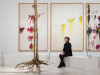A woman is seated on a stark white bench in front of a triptych of three abstract painting while looking at a tree.