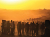 A photograph of a throng of people walking on a dusty landscape at sunset