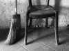 A black and white photograph of the bottom half of a broom and a chair against a rough-hewn wall