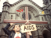 A man in a leather jacket carries a cross with a sign hanging around it that reads, "AIDS," while standing in front of a church