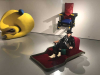 A photograph of Giannina Braschi sitting on a red reclining chair in a gallery space with other unusual furniture displayed
