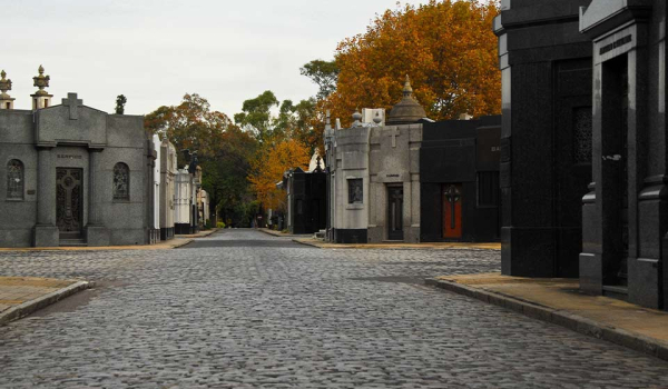 A photograph of a small-stone paved road, lined by ornate mausoleums, inside a tidy cemetary