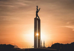 A photograph of a statue of a humanoid figure high on a tower. The tower is in the halo of a sunrise or sunset