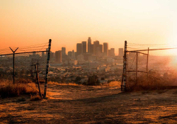 A photograph of a city from a distance, as seen through a ruined chain link fence. An orange haze sits over the city.