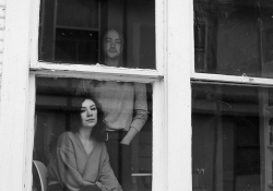 A black and white photograph of a young couple, seen from outside somberly looking at the photographer out a bay window