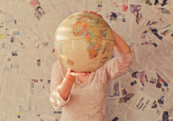 A woman stands in front of a wall covered in newspaper articles, holding up a tan colored globe that covers her face