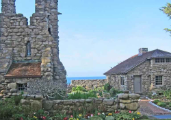A rugged tower rises to the left of a stone house, both of which sit at the edge of the ocean