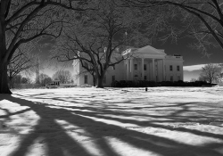 A black and white photo of the White House, seen with long tree shadows lain ominously across the snowy ground