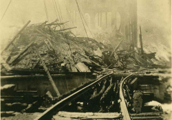 A sepia-toned photograph of a set of railroad tracks, which have been destroyed, with a city in rubble in the background