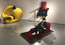 A photograph of Giannina Braschi sitting on a red reclining chair in a gallery space with other unusual furniture displayed