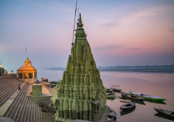 A tall stone temple dominates the shoreline of a wide river, bathed in the colors of sunset